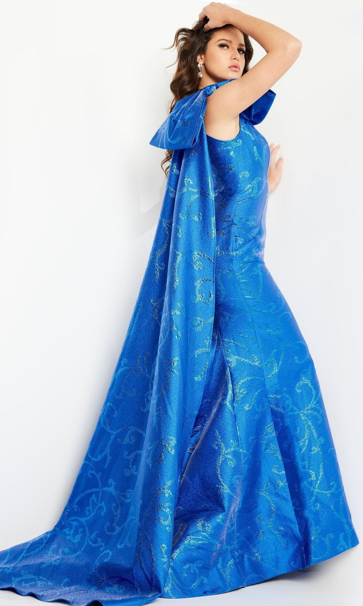 A woman models the Jovani One-Shoulder-Bow Long Formal Dress 23742—a bright blue, textured evening gown featuring a dramatic cape—against a white background.